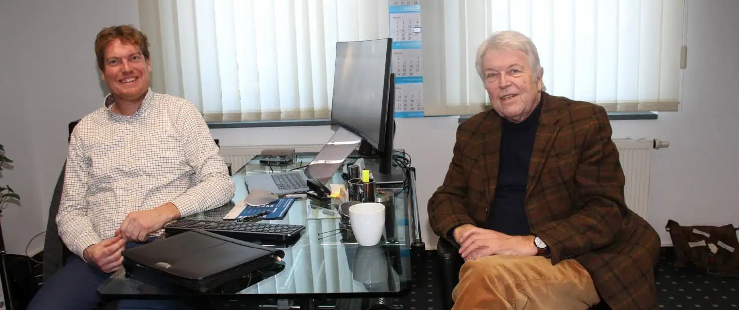 Maximal 100 Zeichen: Zwei Männer sitzen in einem Büro, lächeln und arbeiten am Computer.